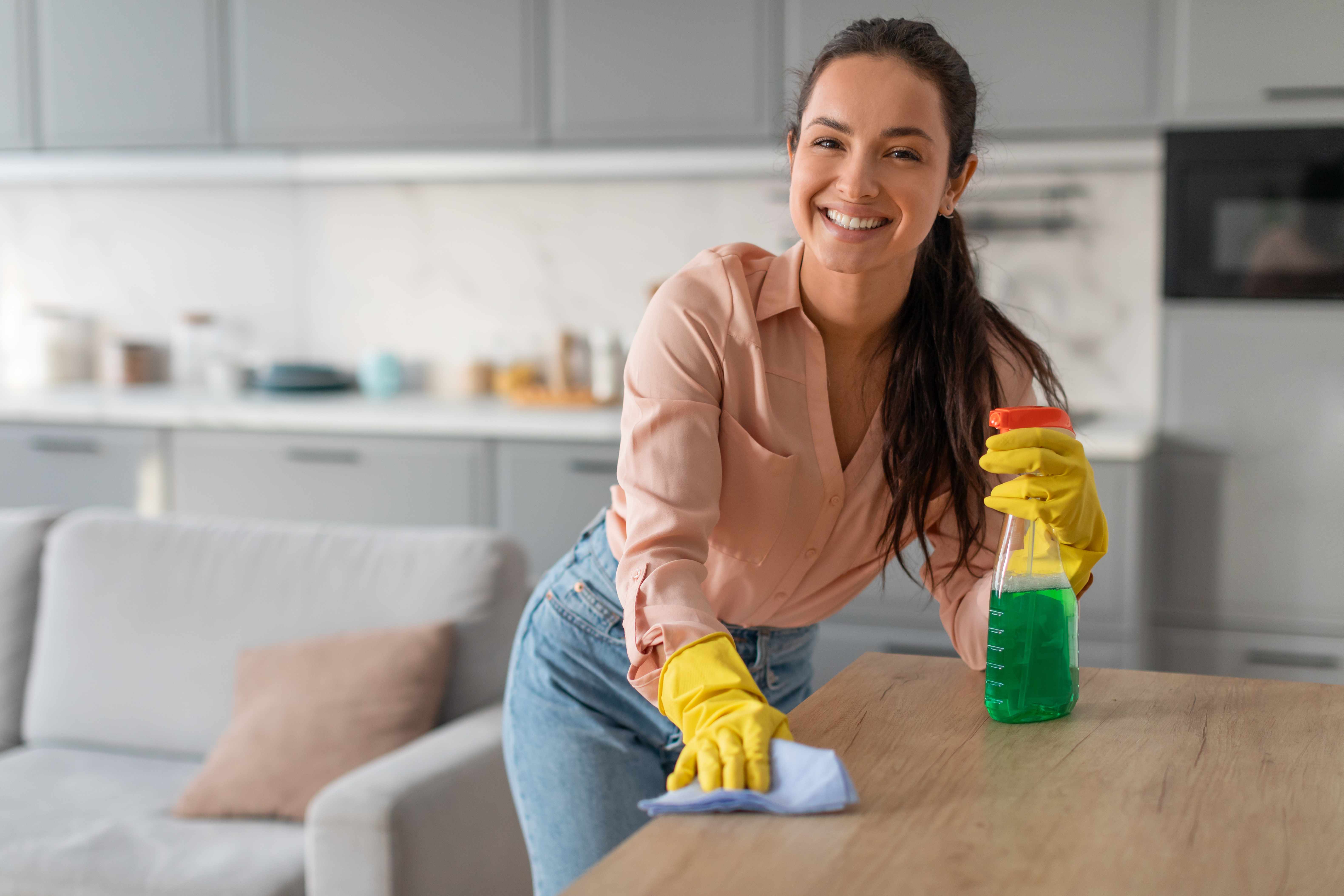 Smiling professional cleaner wiping down table with cleaning supplies in modern kitchen